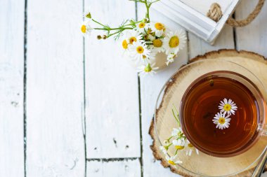 Bir fincan papatya çayı ve beyaz tahta üzerinde papatya çiçekleri olan bitki çayı. Yakınlarda bir vazo beyaz çiçek duruyor. Boşluğu kopyala