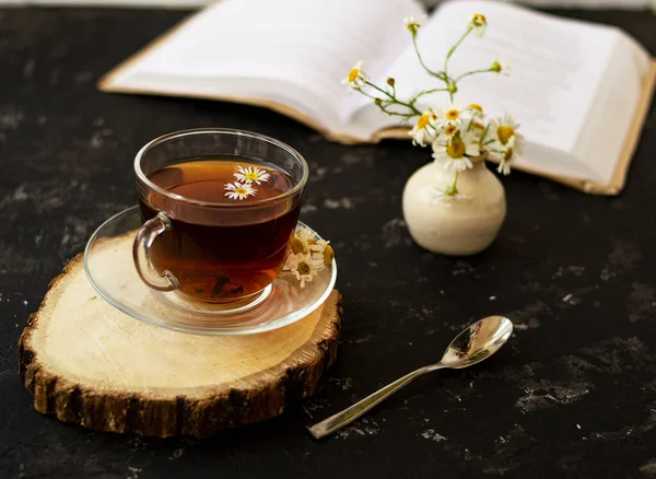 English tea with chamomile stands on a wooden frame and a teaspoon and an open book for reading lies on a black background. Nearby is a white vase with summer chamomile flowers.