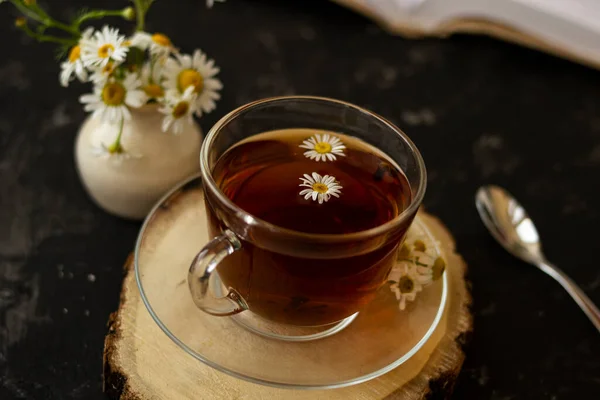 English tea with chamomile stands on a wooden frame and a teaspoon and an open book for reading lies on a black background. Nearby is a white vase with summer chamomile flowers.