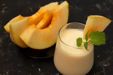 Siyah arka planda kavunlu milkshake. Olgun dilimlenmiş kavun yakınlarda yatıyor. Meyve suyu bardağı nane filizi ve kavun kırıntılarıyla süslenmiş. Yaz sağlıklı gıda konsepti.