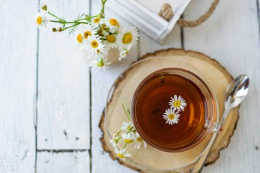 Bir fincan papatya çayı ve beyaz tahta üzerinde papatya çiçekleri olan bitki çayı. Yakınlarda bir vazo beyaz çiçek duruyor. Boşluğu kopyala