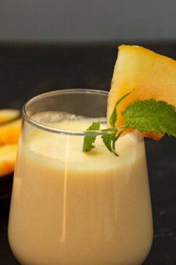 Siyah arka planda kavunlu milkshake. Olgun dilimlenmiş kavun yakınlarda yatıyor. Meyve suyu bardağı nane filizi ve kavun kırıntılarıyla süslenmiş. Yaz sağlıklı gıda konsepti.