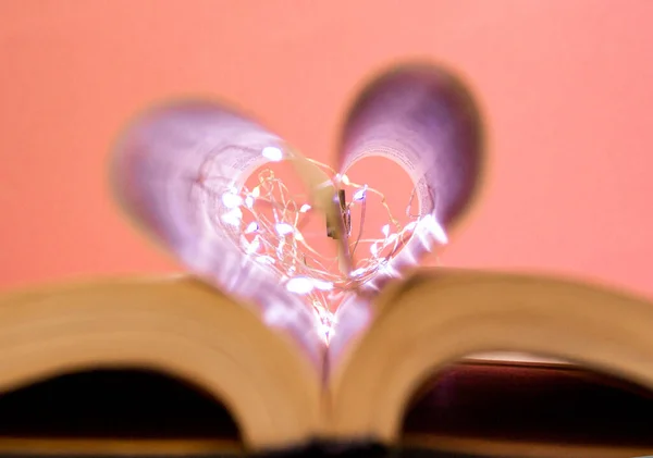 Open book with heart shaped sheets decorated with bright garlands of lights and highlights in the background, romantic love symbol, macro shot. Pink background.