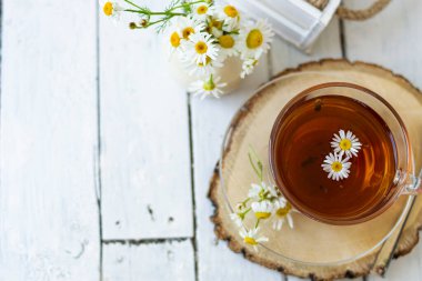 Bir fincan papatya çayı ve beyaz tahta üzerinde papatya çiçekleri olan bitki çayı. Yakınlarda bir vazo beyaz çiçek duruyor. Boşluğu kopyala