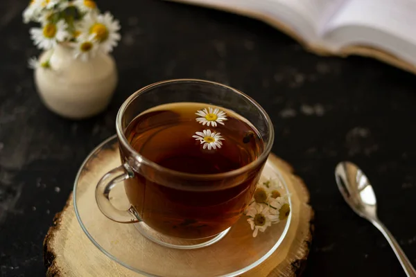 English tea with chamomile stands on a wooden frame and a teaspoon and an open book for reading lies on a black background. Nearby is a white vase with summer chamomile flowers.