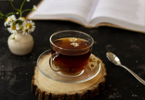 English tea with chamomile stands on a wooden frame and a teaspoon and an open book for reading lies on a black background. Nearby is a white vase with summer chamomile flowers.