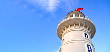 Mavi bir gökyüzüne karşı beyaz bir deniz feneri duruyor. Deniz fenerinin üzerinde kırmızı bir Türk bayrağı dalgalanıyor. Deniz feneri, Türkiye, Alanya.