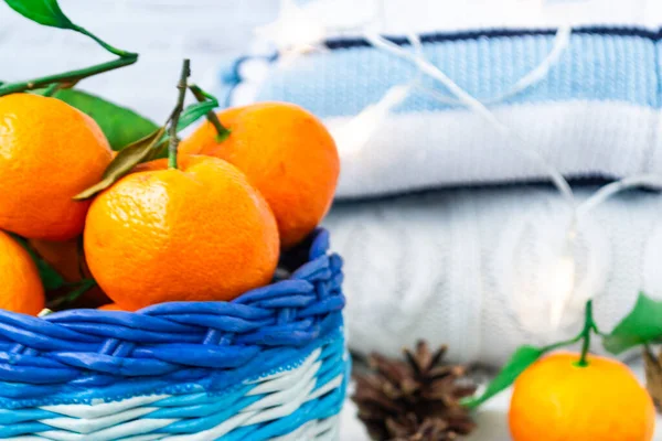 Christmas basket with tangerines, cinnamon sticks and pine cones lie on a wooden white background. Knitted jumpers stack on top of each other and the garland burns beautifully. Hygge concept.