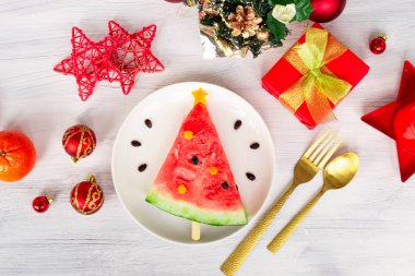 Bir parça karpuz, beyaz tabakta duran bir Noel ağacı gibi süslenir. Yıldızlı, balonlu ve dekorlu beyaz bir arka plan. Çocuklar için kahvaltı fikri. Yeni yıl, Noel yemeği, en iyi manzara. kutlama,