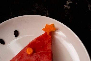 Bir parça karpuz, beyaz tabakta duran bir Noel ağacı gibi süslenir. Betonun altındaki siyah arka planda altın çatallı ve kaşıklı. Çocuklar için kahvaltı fikri. Yeni yıl, karpuzlu Noel yemeği, üst manzara. kutlama.