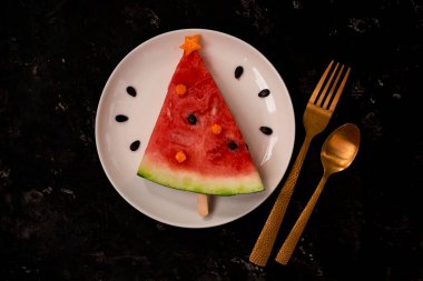 Bir parça karpuz, beyaz tabakta duran bir Noel ağacı gibi süslenir. Betonun altındaki siyah arka planda altın çatallı ve kaşıklı. Çocuklar için kahvaltı fikri. Yeni yıl, karpuzlu Noel yemeği, üst manzara. kutlama.