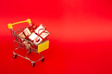 Kırmızı arka planda kırmızı afiş ve fotokopi alanı olan altın hediye kutularının olduğu market sepeti. İnternetten Noel alışverişi. Kış tatili satışları, mevsimlik satışlar, Kara Cuma, Noel, indirimler ve online alışveriş. Xmas satışı.