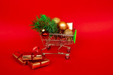 Altın hediye kutuları ve noel baloları olan market sepeti, kırmızı afiş arkaplanda fotokopi alanı olan noel ağacı filizi. Noel alışverişi internetten. Kış tatili indirimleri, mevsimlik satışlar, Kara Cuma, Noel, indirimler.