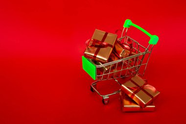 Kırmızı arka planda kırmızı afiş ve fotokopi alanı olan altın hediye kutularının olduğu market sepeti. İnternetten Noel alışverişi. Kış tatili satışları, mevsimlik satışlar, Kara Cuma, Noel, indirimler ve online alışveriş. Xmas satışı.