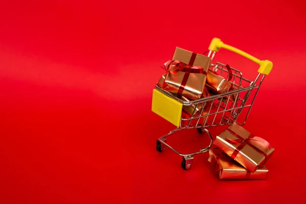 Grocery basket with golden gift boxes with red ribbon on red background banner with copy space. Christmas shopping online. Winter holiday sales, seasonal sales, Black Friday, Christmas, discounts and online shopping. Xmas sale.