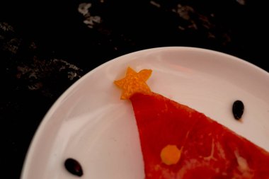 Bir parça karpuz, beyaz tabakta duran bir Noel ağacı gibi süslenir. Betonun altındaki siyah arka planda altın çatallı ve kaşıklı. Çocuklar için kahvaltı fikri. Yeni yıl, karpuzlu Noel yemeği, üst manzara. kutlama.