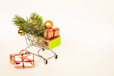 Altın hediye kutuları ve Noel baloları olan market sepeti, arka planda beyaz afişlerle süslenmiş Noel ağacı filizi. Xmas indirimi. İnternetten Noel alışverişi. Kış tatili indirimleri, mevsimlik satışlar, Kara Cuma, Noel, indirim.