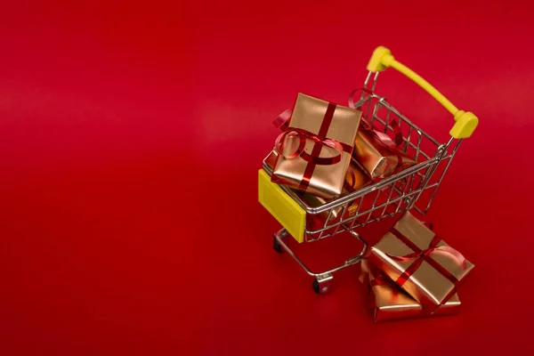 Grocery basket with golden gift boxes with red ribbon on red background banner with copy space. Christmas shopping online. Winter holiday sales, seasonal sales, Black Friday, Christmas, discounts and online shopping. Xmas sale.
