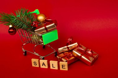 İçinde altın hediye kutuları ve Noel topları olan market sepeti, Noel ağacı filizi ve kırmızı arka planda fotokopi alanı olan İngilizce harflerle satılıyor. Xmas indirimi. İnternetten Noel alışverişi. Kış
