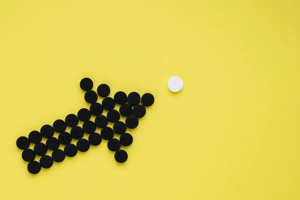 Medical activated carbon in the form of an arrow pointing to a white pill on a yellow background. Top view, copy space.