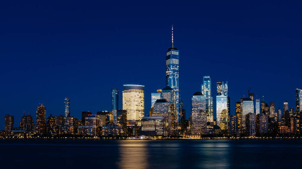 Night view of skyline of downtown Manhattan over Hudson River under dark blue sky, viewed from New Jersey, in New York City, USA