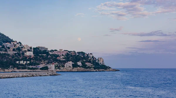 View of Lighthouse of Nice and houses on hills by blue sea, under the moon, in Nice, France