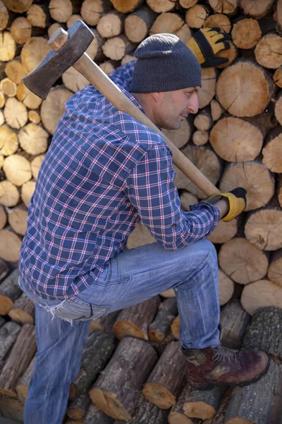 Un uomo con un'ascia industriale. Ax in mano. Un uomo forte tiene in mano un'ascia sullo sfondo di motoseghe e legna da ardere. Uomo forte boscaiolo con un'ascia in mano. motosega da vicino. — Foto Stock