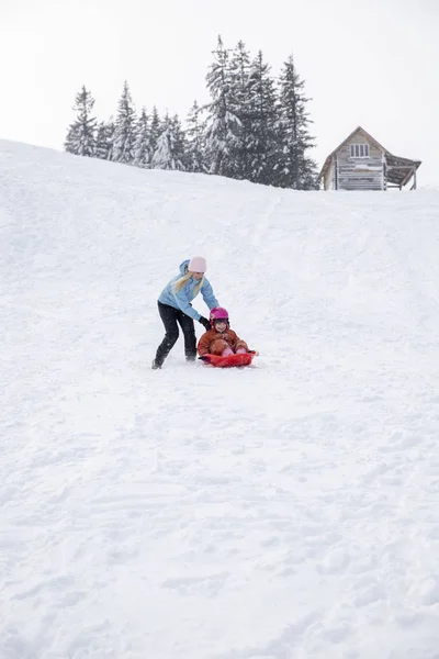 Moeder en dochter rijden op een slee vanaf een dia sneeuw. Rijden vanaf een heuvel van de sneeuw op een slee. Familie slee rijdt, slee rijdt, Winterpret, sneeuw. — Stockfoto