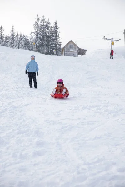 Moeder en dochter rijden op een slee vanaf een dia sneeuw. Rijden vanaf een heuvel van de sneeuw op een slee. Familie slee rijdt, slee rijdt, Winterpret, sneeuw. — Stockfoto