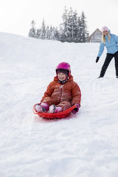 Mamá y su hija viajan en trineo desde un tobogán de nieve. Cabalga desde una colina de nieve en un trineo. Paseos en trineo, diversión de invierno, nieve, paseos en trineo familiar . — Foto de Stock