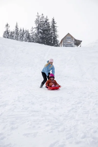 Moeder en dochter rijden op een slee vanaf een dia sneeuw. Rijden vanaf een heuvel van de sneeuw op een slee. Familie slee rijdt, slee rijdt, Winterpret, sneeuw. — Stockfoto