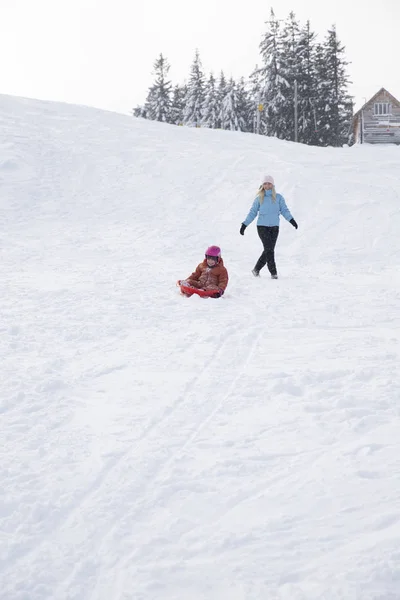 Μαμά και κόρη βόλτα σε ένα έλκηθρο από μια διαφάνεια χιόνι. Βόλτα από ένα hill χιονιού σε ένα έλκηθρο. Βόλτες με έλκηθρα, βόλτες με έλκηθρα οικογενειακή διασκέδαση χειμώνα, χιόνι. — Φωτογραφία Αρχείου