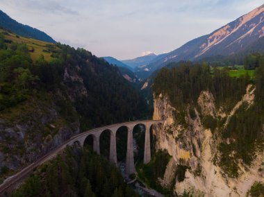 İsviçre Alpleri 'ndeki eğri bir raydaki taş bir köprüden geçen kırmızı dar gösterge..