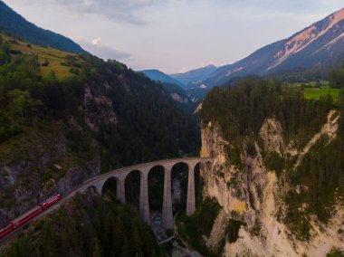 İsviçre Alpleri 'ndeki eğri bir raydaki taş bir köprüden geçen kırmızı dar gösterge..