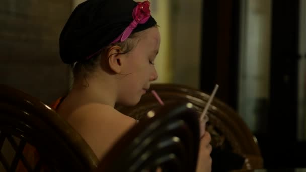 Маленькая девочка в плавательных очках пьет коктейль через соломинку в кафе у бассейна. 4K — стоковое видео