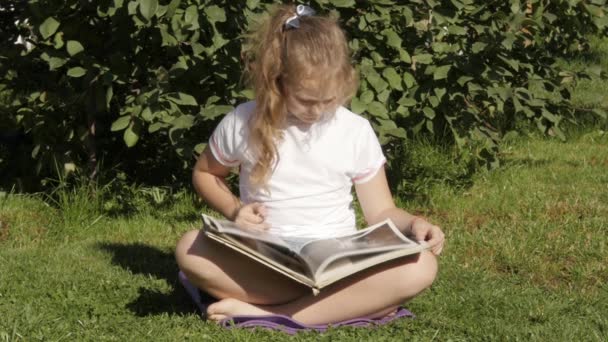 Krásné dospívající dívka sedí na trávě v létě park a čte knihu. Zpomalený pohyb — Stock video