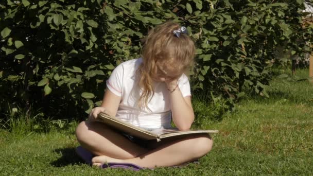 Красивая девочка-подросток сидит на траве в летнем парке и читает книгу. замедленное движение — стоковое видео