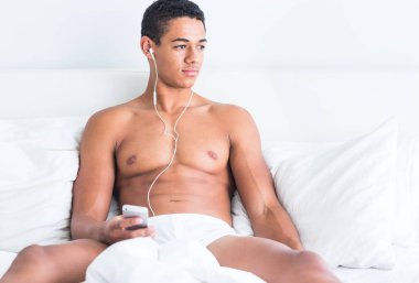 Müzik yaptığı telefon dinlemek bir beyaz yatakta seksi üstsüz siyah Afrika adam oturuyor. Gevşeme ve kurtarma.