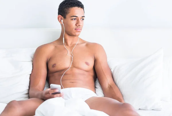 Sexy topless black African man sitting on a white bed listening to music on his phone. Relaxation and recovery.