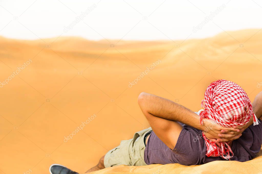 Backshot of a Caucasian man wearing a Middle Eastern headwear and reclined in the desert sand during vacation