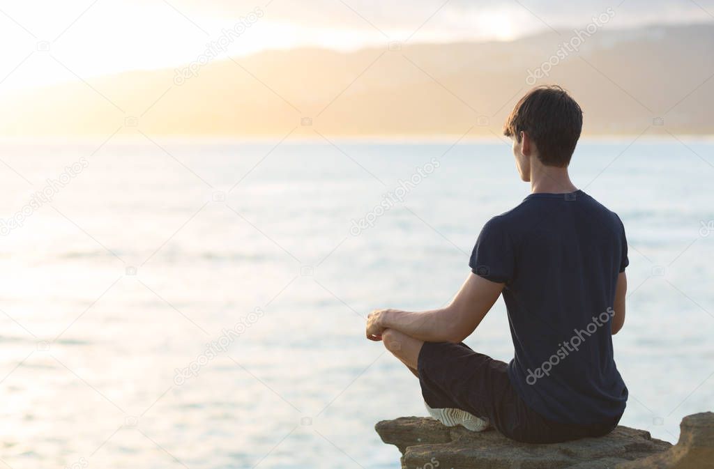 A young man watching the ocean sunset while doing a meditation yoga pose. Relaxation and reflection.
