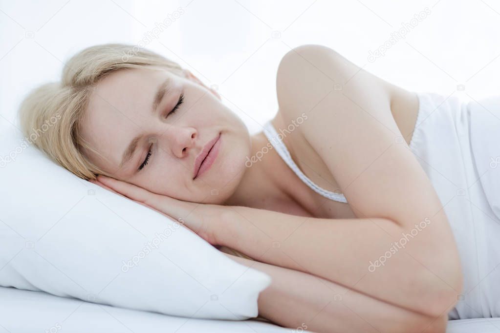 A beautiful blonde woman sleeping on a white soft pillow and bed at home. Relaxation and rejuvenation.