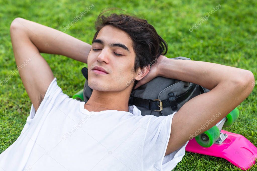 Young college male student taking a nap on the grass outside, with his backpack and skateboard. 