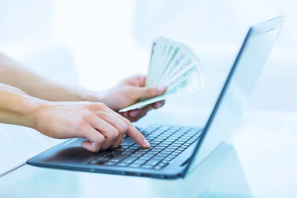 Close-up of hands shopping/paying online using laptop while holding US dollars. Finance management.