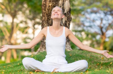 Mutlu genç bir kadın kendini huzurlu ve minnettar hissediyor. Kollarını açıp gökyüzüne bakarken parktaki bir ağacın altında çimlerin üzerinde oturuyor..