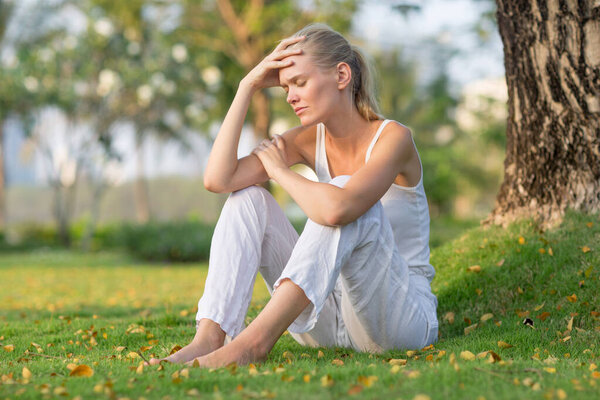 Sad woman sitting alone on the grass at the park with hand on forehead. Feeling bad and depressed.