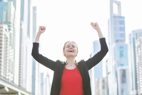 Business and career success - Happy young businesswoman celebrating work achievements with both hands up against a modern city\'s high-rise buildings in the background.