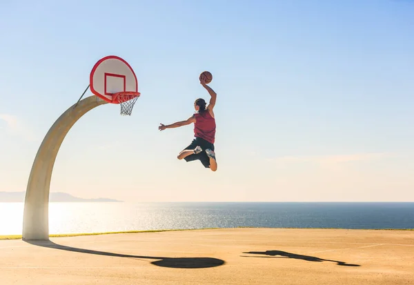 Basketbolcu havada zıplıyor ve arka planda güzel bir okyanus manzarası olan sokak topu sahasında inanılmaz enerjik bir smaç basıyor. Spor, motivasyon ve yükseklik.