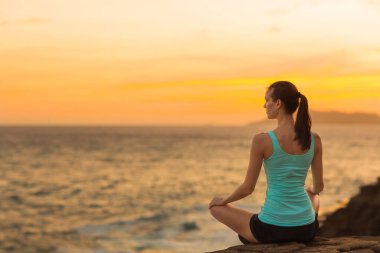 Sakin ve rahat bir kadın, turuncu bir günbatımı gökyüzü boyunca güzel bir okyanus manzarası olan sahilde huzur içinde meditasyon yapıyor. Sağlık, yoga ve uyum.