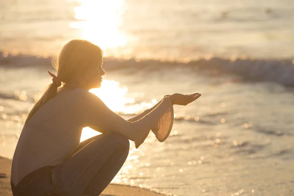 Calm and peaceful young woman sitting alone on the beach sand during a beautiful sunset reflecting on water. Relaxing and self.